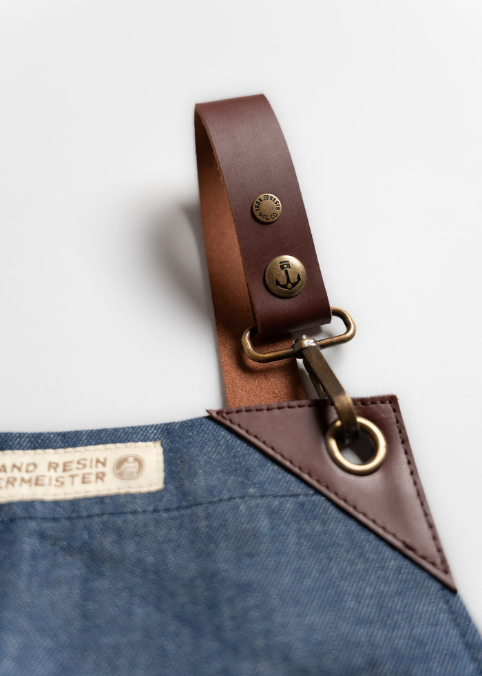 Close-up of a denim apron with a brown leather strap and brass anchor button. Label reads "Iron and Resin Werkmeister."