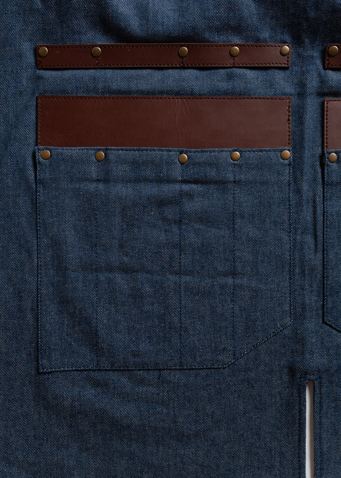 Denim apron with brown leather accents and rivets, featuring large front pockets. Durable and stylish design for work or craft.