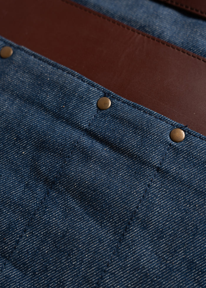 Close-up of blue denim fabric with brown leather trim and metal rivets, showcasing texture and craftsmanship details.