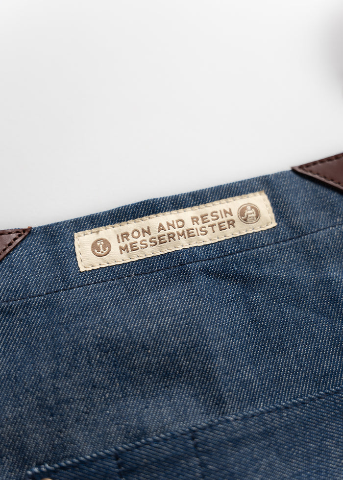 Close-up of a denim apron with a label reading "Iron and Resin Messermeister," featuring leather accents on a white background.
