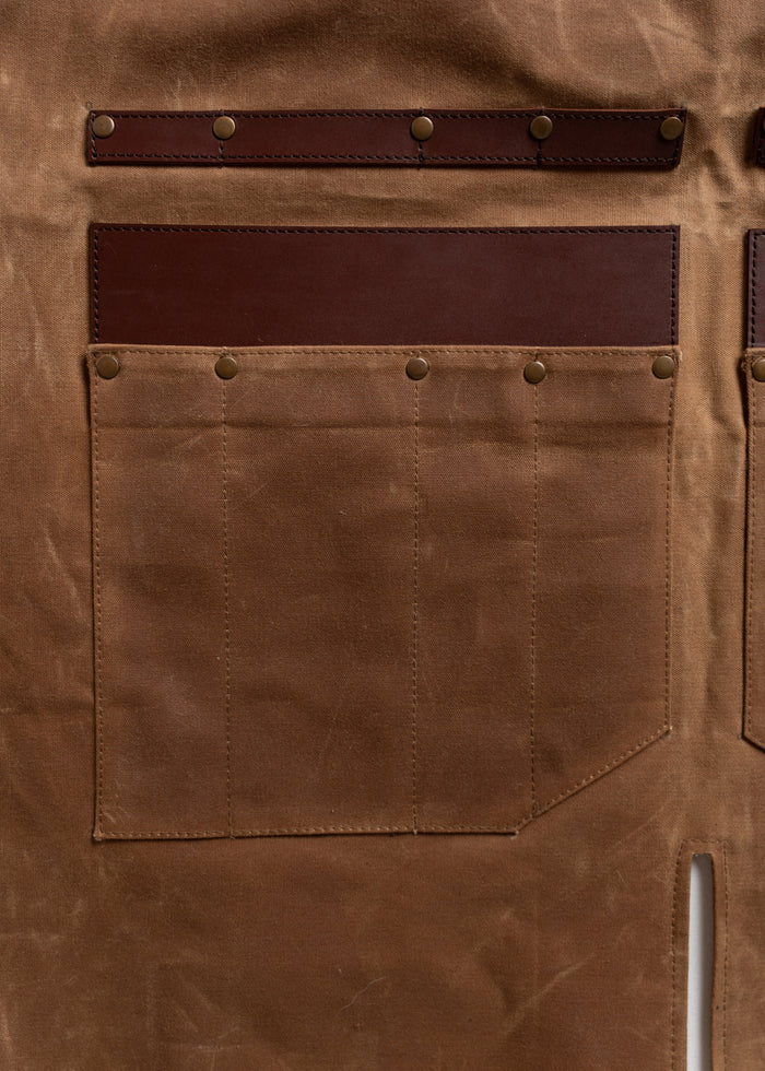 Brown waxed canvas apron with leather straps and brass rivets, featuring multiple pockets for tools and accessories.