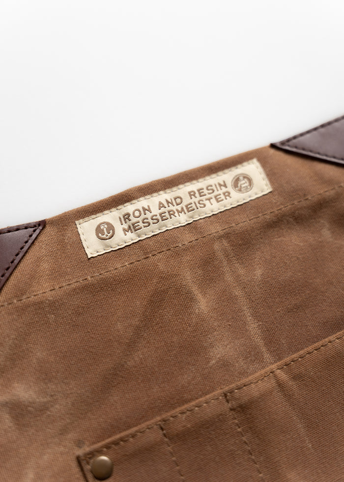 Brown fabric apron with "Iron and Resin Messermeister" label, featuring leather corners and a riveted pocket for durability.