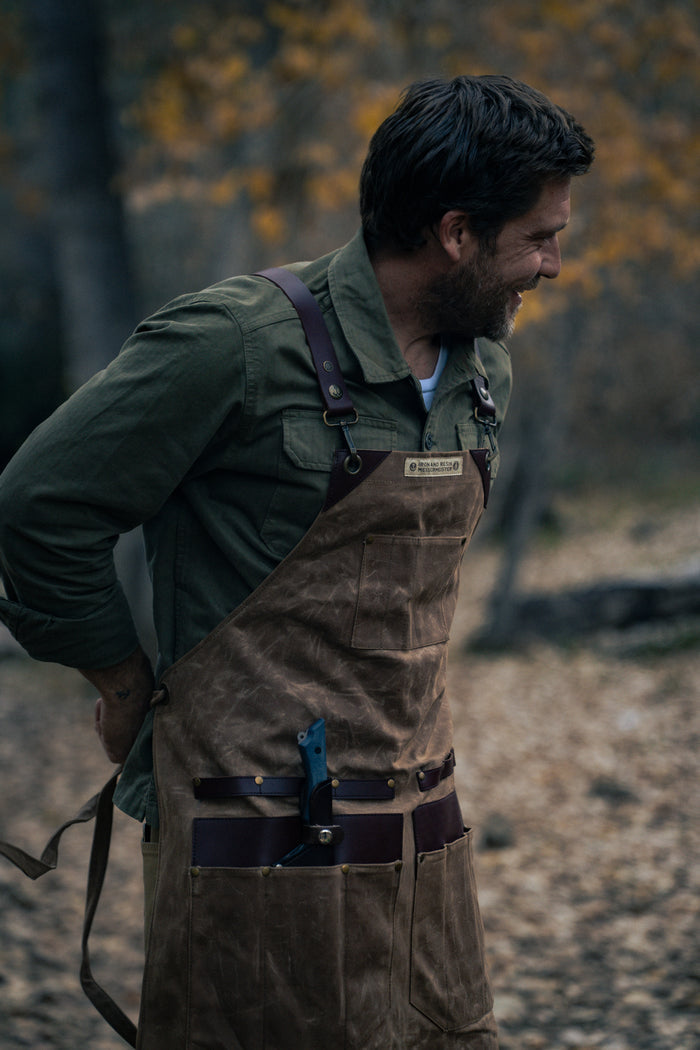 Man in green shirt and brown apron with knife in pocket, standing outdoors with autumn leaves in background, smiling to the side.