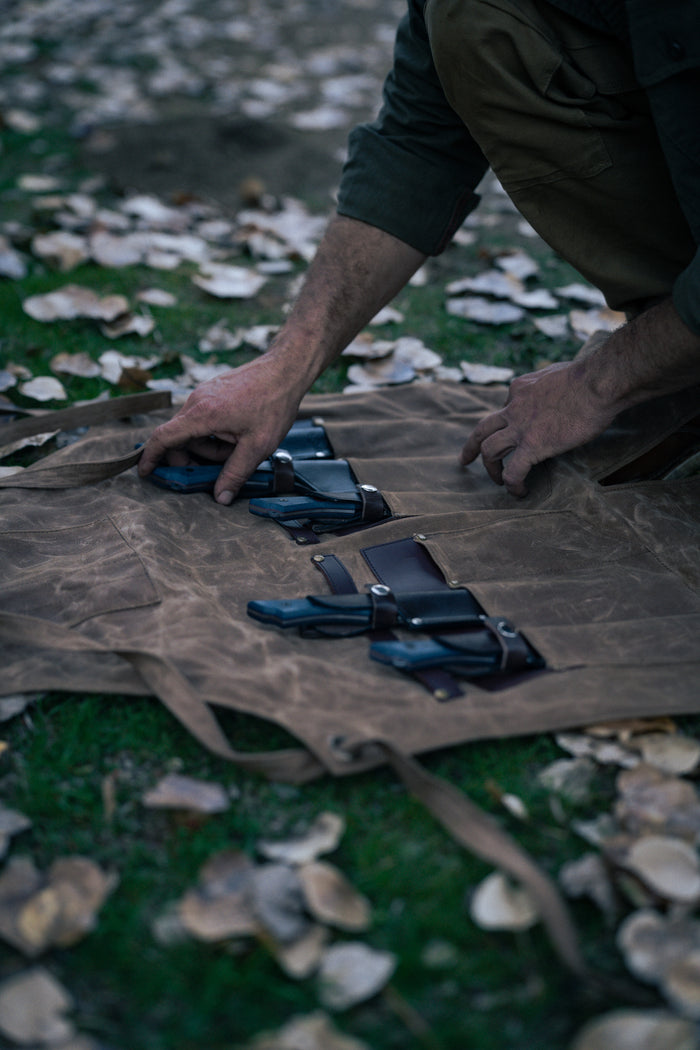 A person arranges knives on a brown canvas roll outdoors, surrounded by fallen leaves, highlighting a rustic, natural setting.