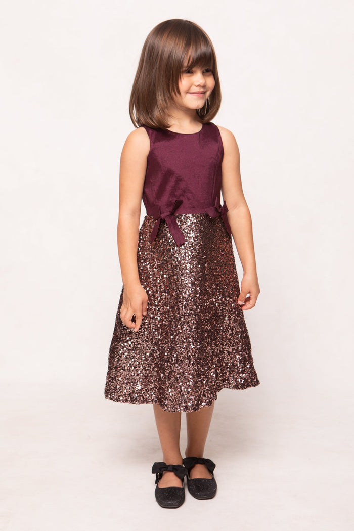 A young girl with straight brown hair is wearing the Ivy City Co Mini Alexis Dress in Wine—a sleeveless burgundy festive dress with a sparkly sequin skirt. She pairs it with black shoes featuring bows and stands against a plain white background, smiling slightly.