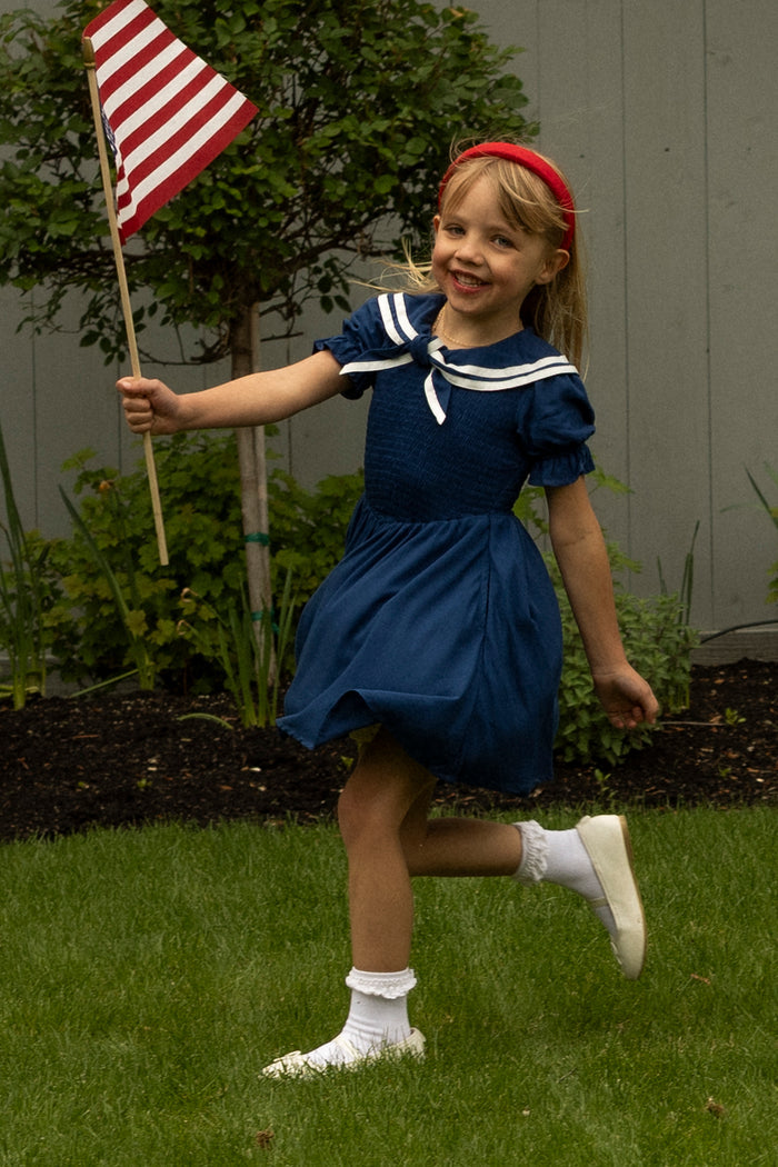A young girl wears the Ivy City Co Mini Alice Dress in Navy, paired with white socks and shoes, as she waves an American flag and skips joyfully on a grassy lawn. She has a red headband; green plants and a gray fence are in the background.