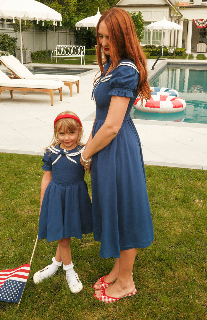 A woman and a young girl stand on grass by a pool, both wearing Ivy City Co Mini Alice Dresses in Navy with white trim. The girl holds an American flag and wears a red headband. Pool floats and lounge chairs are in the background.