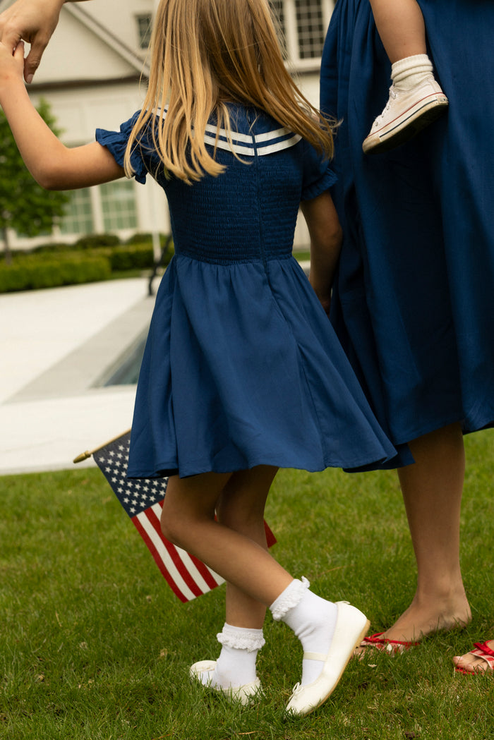 A young girl in the Ivy City Co Mini Alice Dress in Navy holds hands with an adult carrying a child. She waves an American flag on the grass near a house, creating a classic matching Mom and Me moment.
