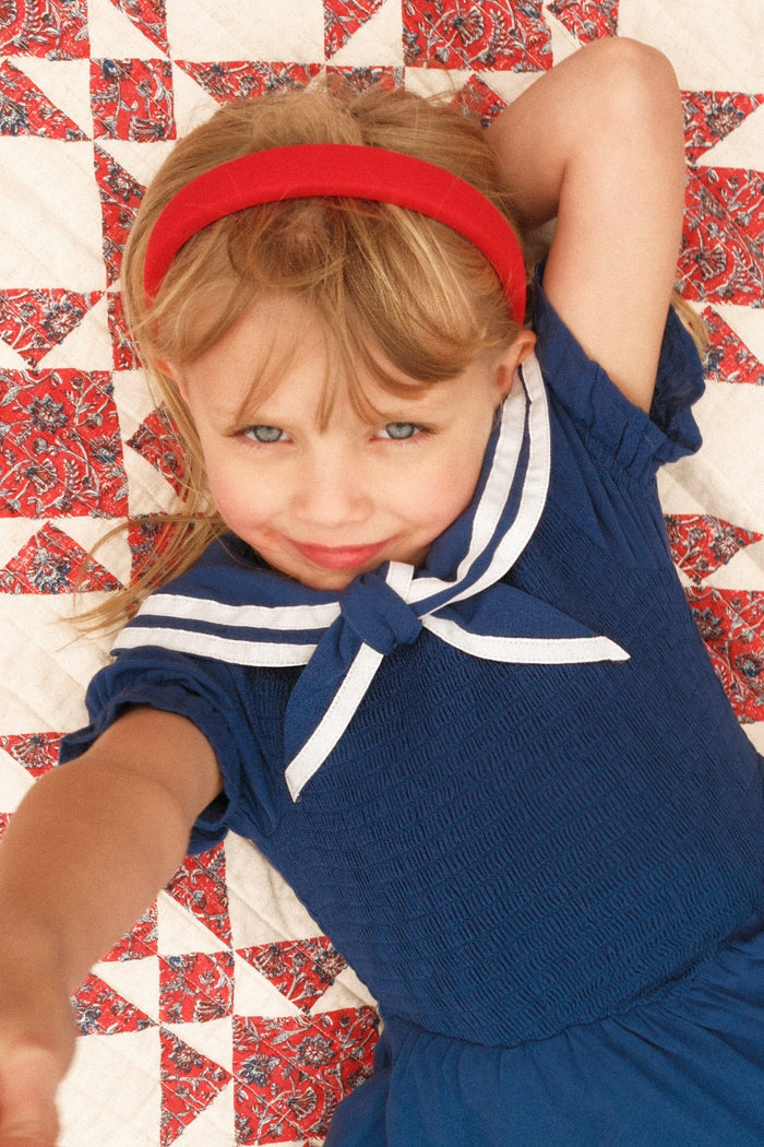 A young girl with blonde hair and blue eyes, wearing the Ivy City Co Mini Alice Dress in Navy with a sailor collar and a red headband, smiles and reaches forward while lying on a red, white, and blue patterned quilt.