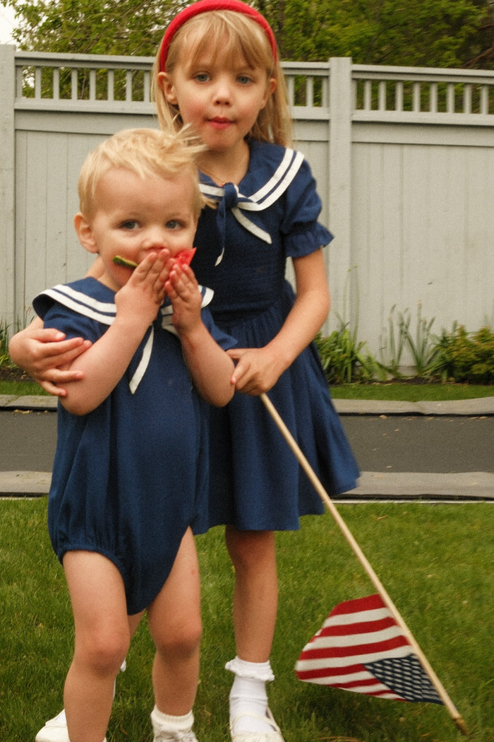 Two young children stand on grass by a fence in matching navy outfits with white sailor collars. The older wears the Ivy City Co Mini Alice Dress in Navy and holds an American flag, while the younger covers their mouth with their hands.