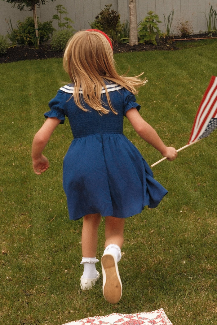 A young girl in the Ivy City Co Mini Alice Dress in Navy runs across a grassy yard holding an American flag, her back to the camera. Trees and a fence can be seen in the background.