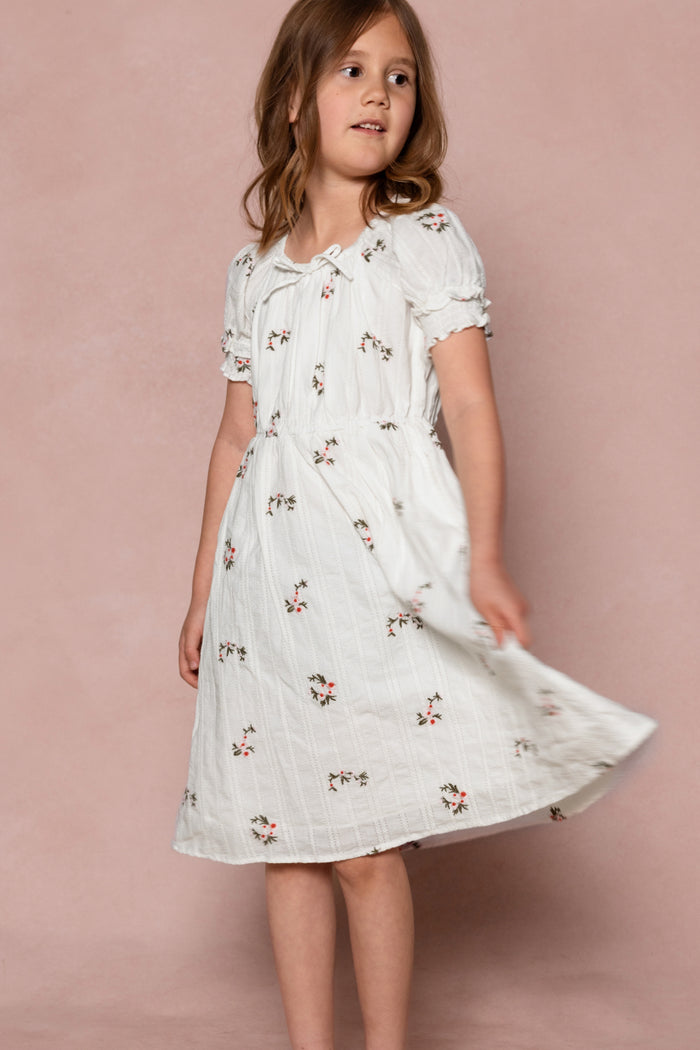 A young girl with light brown hair wears the Ivy City Co Mini Jessie Dress in Ditsy Embroidery, standing against a pink background and twirling as she looks slightly to the side.