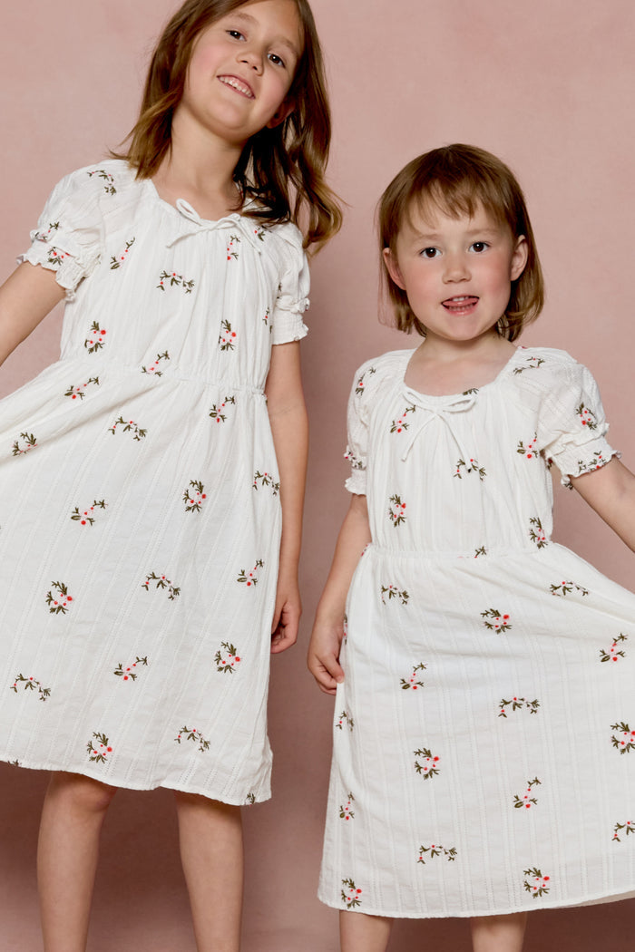 Two young children wear Ivy City Co’s Mini Jessie Dress in Ditsy Embroidery, standing side by side against a pink background, smiling as they hold out the sides of their white, floral-patterned embroidered cotton dresses.