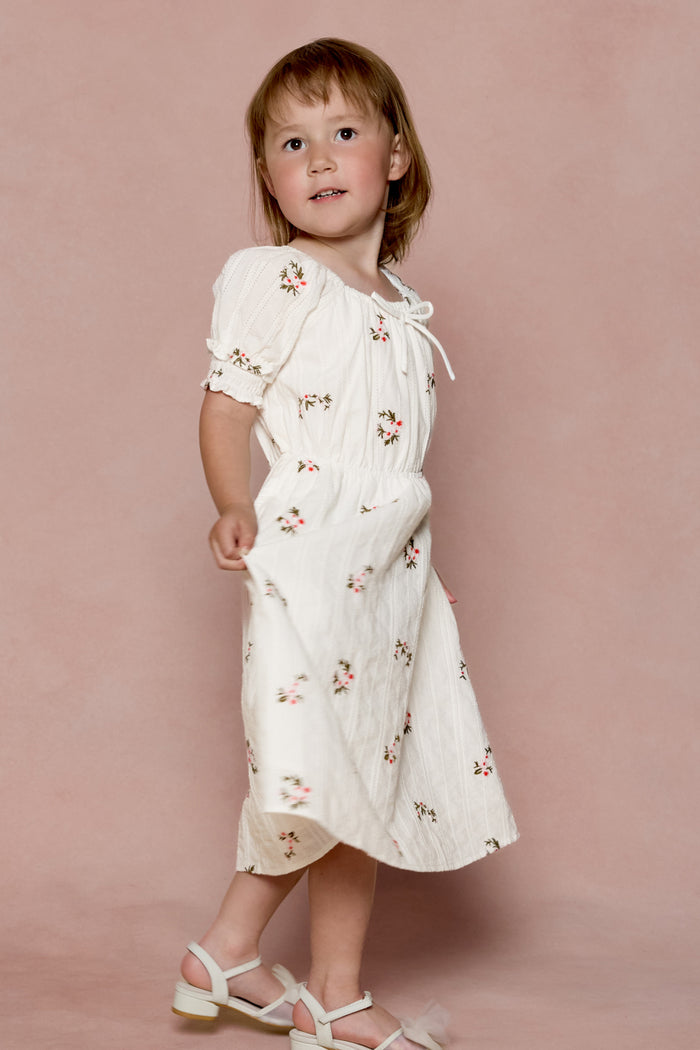 A young child wearing the Ivy City Co Mini Jessie Dress in Ditsy Embroidery and white sandals stands against a soft pink background, gazing slightly upward.