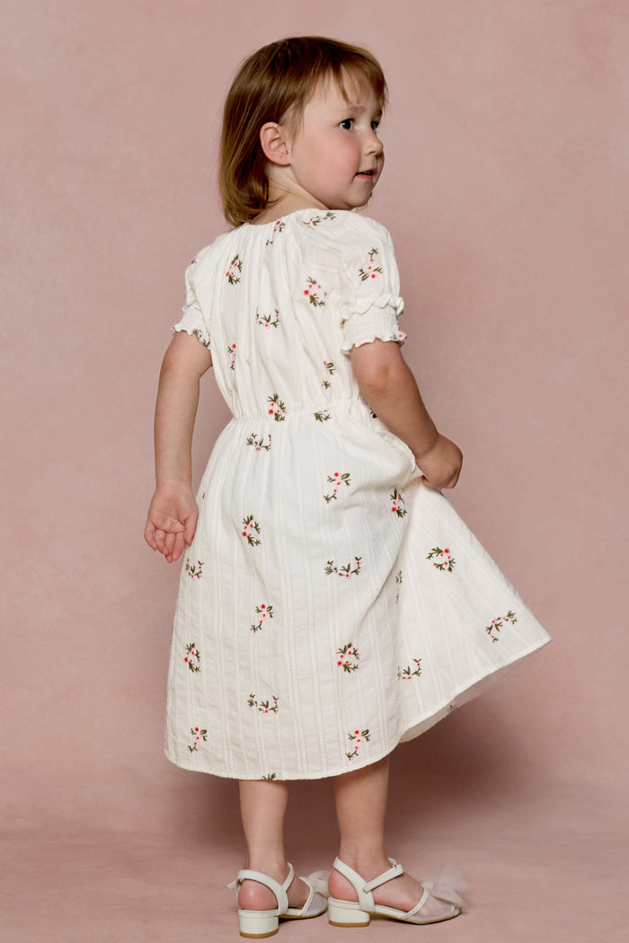 A young child with light brown hair stands sideways, looking over their shoulder in the Mini Jessie Dress in Ditsy Embroidery by Ivy City Co— a cream-colored embroidered cotton dress— paired with white sandals against a soft pink backdrop.