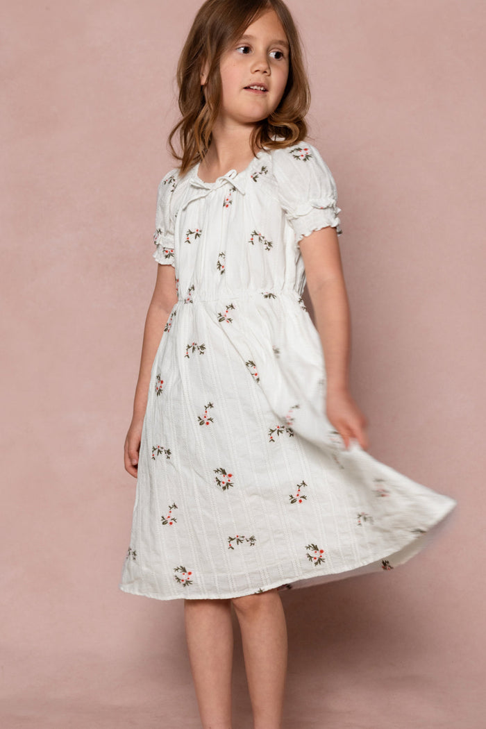 A young girl twirls gently against a light pink background, wearing the Ivy City Co Mini Jessie Dress in Ditsy Embroidery—featuring short sleeves and delicate small floral prints.