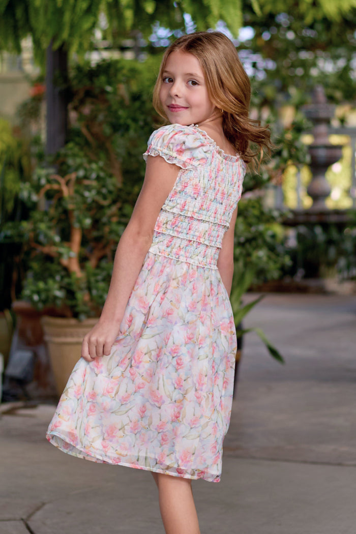 A young girl with light brown hair smiles over her shoulder while wearing the Ivy City Co Mini Kyla Dress in Watercolor Orchids, standing outdoors among green plants.