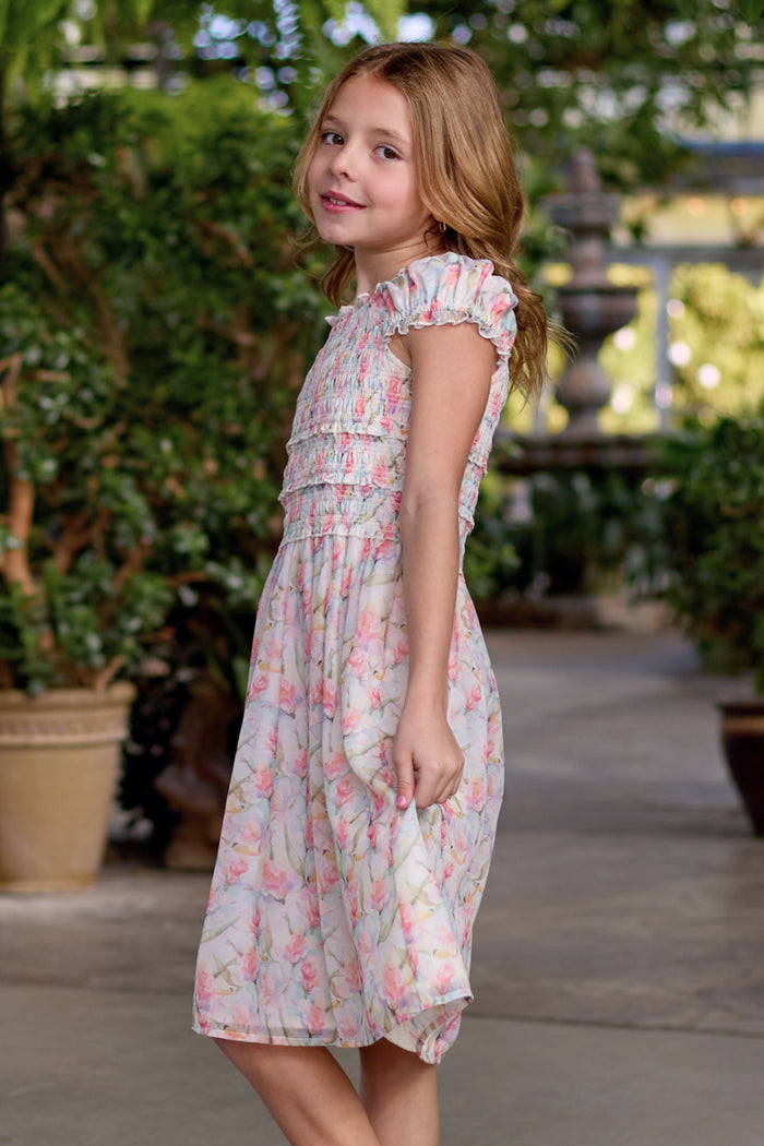 A young girl with wavy light brown hair wears the Mini Kyla Dress in Watercolor Orchids by Ivy City Co, looking back over her shoulder, surrounded by green plants and potted flowers in a charming garden setting.