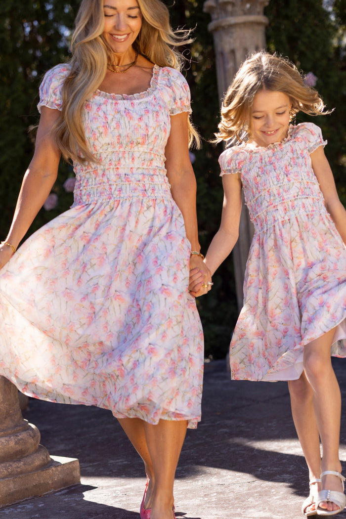 A woman and a young girl hold hands and smile outdoors on a sunny day, both wearing Ivy City Co's Mini Kyla Dress in Watercolor Orchids.