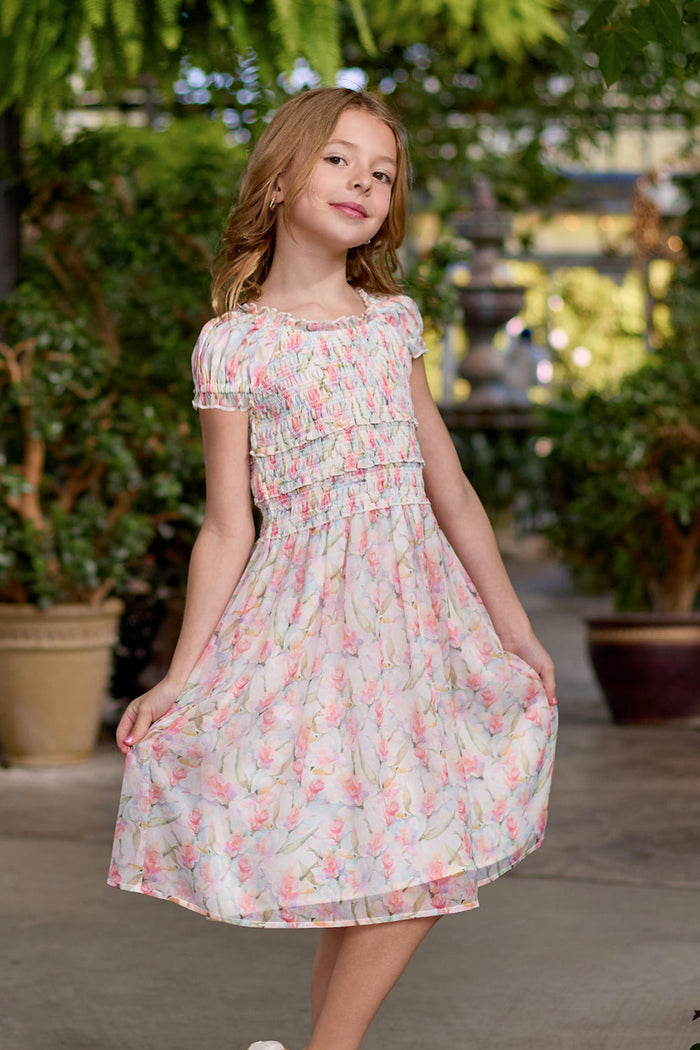 A young girl wearing the Ivy City Co Mini Kyla Dress in Watercolor Orchids stands outdoors, gently holding the sides of her skirt. She is surrounded by lush green plants and potted flowers, bathed in soft natural light.