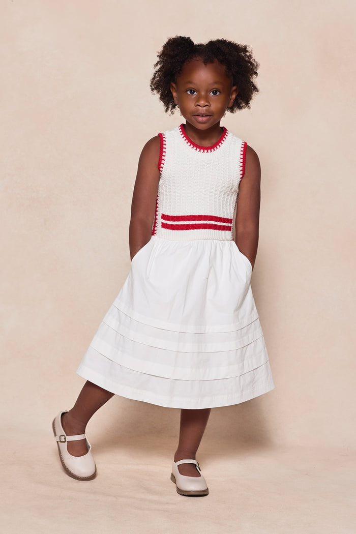 A young girl wears the Mini Lisbon Dress in Ivory & Firework Red by Ivy City Co—a sleeveless white dress with red accents, perfect for the Fourth of July. She stands against a beige background, hands in her pockets, wearing light shoes.