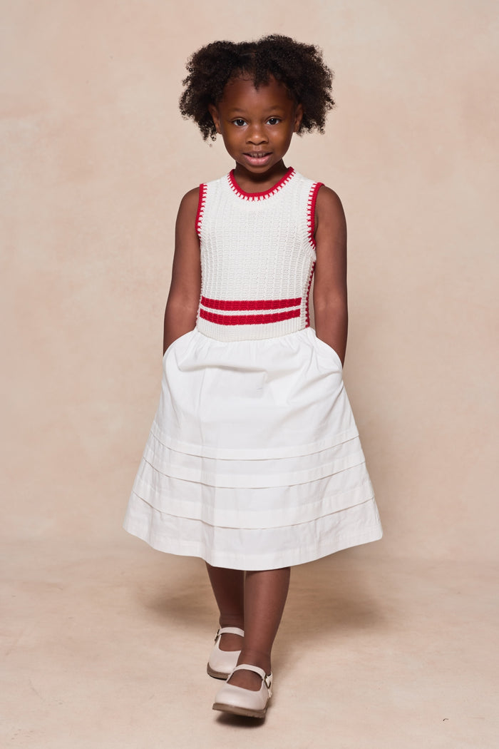 A young girl with curly hair stands with her hands in the pockets of her Ivy City Co Mini Lisbon Dress in Ivory & Firework Red, featuring a sleeveless top and red trim, paired with white shoes against a light beige background.
