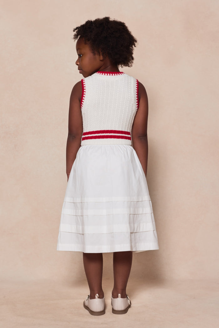 A young girl with curly hair stands facing away, wearing the Ivy City Co Mini Lisbon Dress in Ivory & Firework Red—sleeveless with red trim and stripes near the hem—and white shoes against a beige background. Perfect for Mom and Me matching moments.