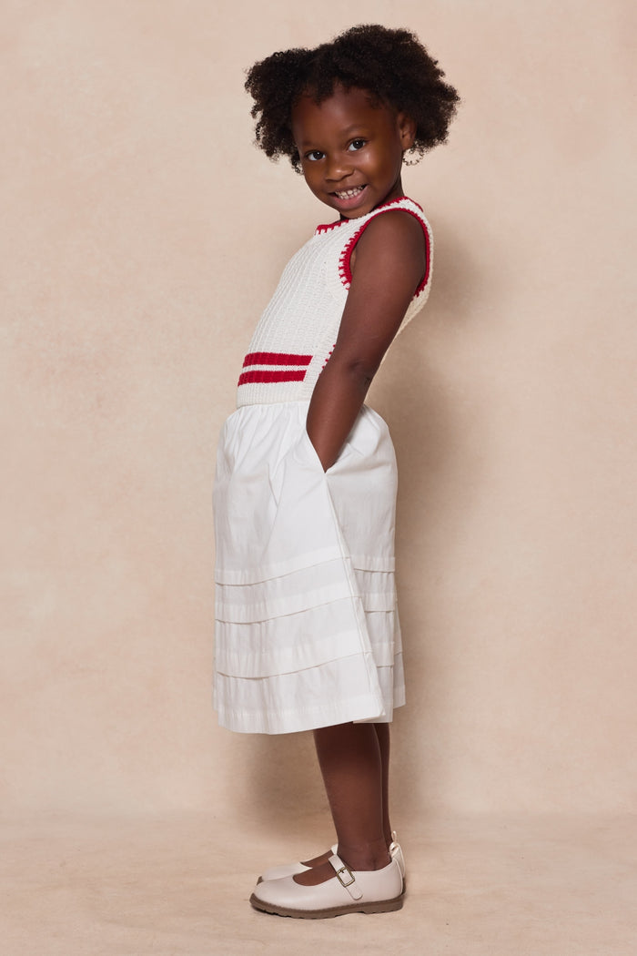 A young girl with curly hair smiles, wearing Ivy City Co's Mini Lisbon Dress in Ivory & Firework Red and beige Mary Jane shoes, standing sideways with her hands in her pockets against a light beige background.