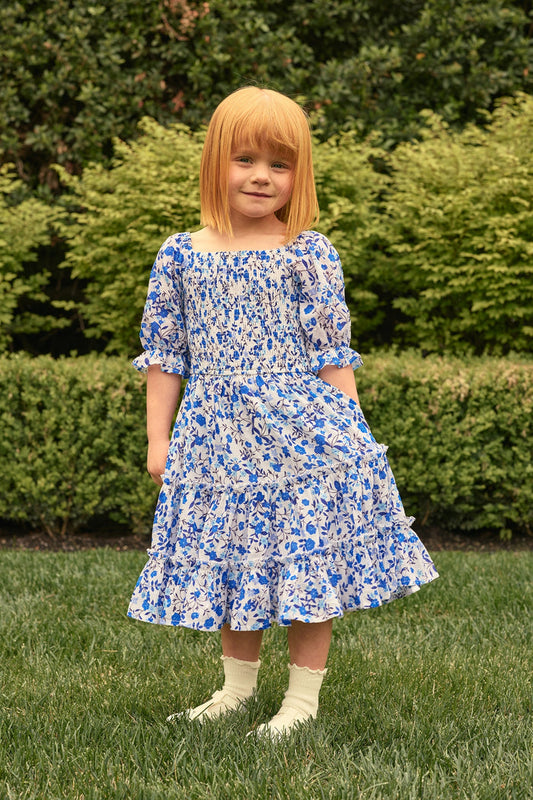 A young girl wears the Ivy City Co Mini Madeline Dress in Ditsy Cobalt Floral, paired with white socks and cream shoes, standing on grass with lush greenery—ideal for a charming Mom and Me matching moment.