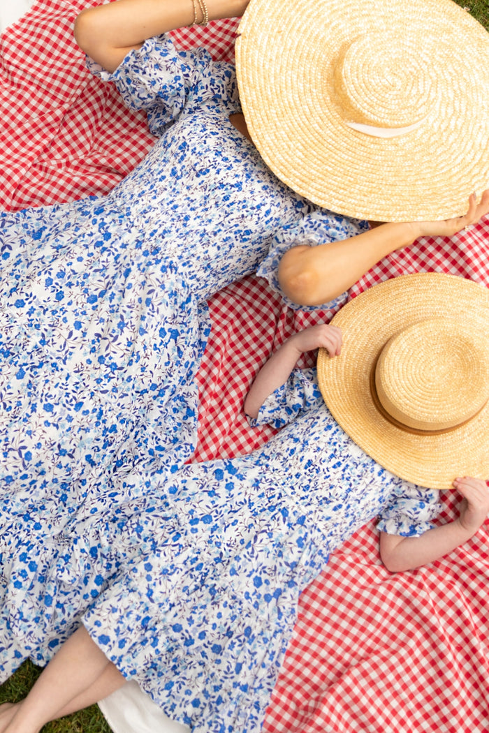 Two people wear Ivy City Co’s Mini Madeline Dress in Ditsy Cobalt Floral, lying side by side on a red and white checkered picnic blanket with large straw hats partially covering their faces.