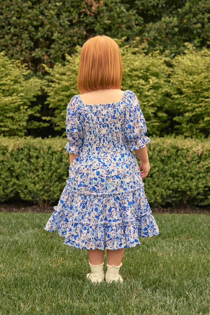 A young girl with straight red hair wears the Ivy City Co Mini Madeline Dress in Ditsy Cobalt Floral, paired with white socks and shoes, standing on grass and facing away from the camera, with green bushes in the background.