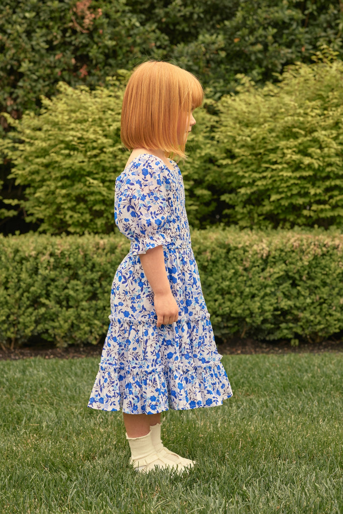 A young child with straight red hair, wearing the Ivy City Co Mini Madeline Dress in Ditsy Cobalt Floral and white socks, stands in profile on a grassy lawn with green bushes in the background.