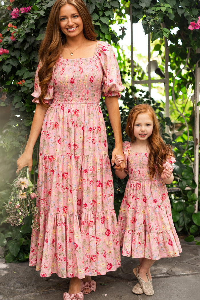 A woman and young girl, possibly mother and daughter, smile hand-in-hand in lush greenery, both in matching Ivy City Co dresses. The girl wears the Mini Madeline Dress in Pink Vintage Poppies.