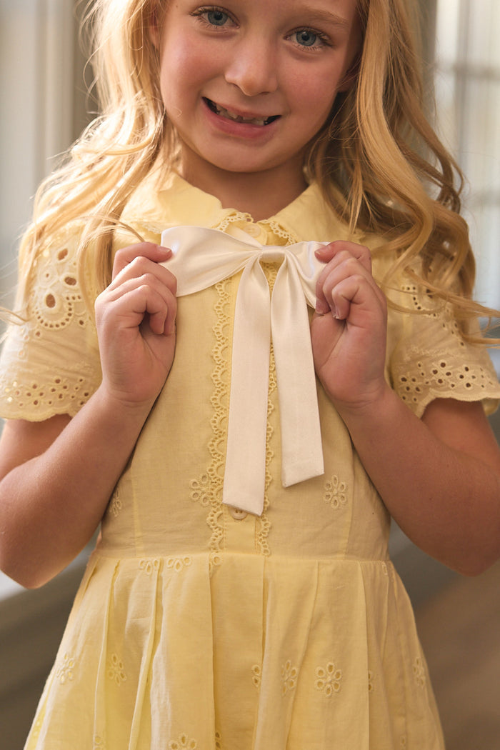 A young blonde girl wears the Ivy City Co Mini Penny Dress in Butter Yellow Eyelet, holding a white ribbon bow at her collar and smiling softly as gentle window light fills the background.