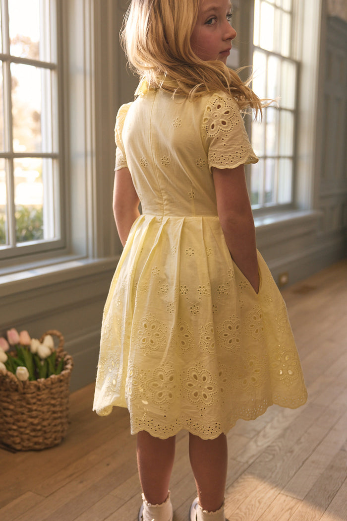 A young girl with blonde hair stands by tall windows in a sunlit room, wearing the Ivy City Co Mini Penny Dress in Butter Yellow Eyelet. A basket of tulips on the floor sets the scene for charming mama and mini matching moments.