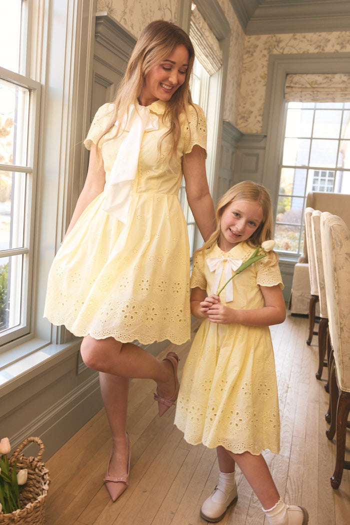 A woman and young girl smile in a sunlit room, both wearing Ivy City Co’s Mini Penny Dress in Butter Yellow Eyelet. The girl holds a tulip, with a basket of flowers on the floor nearby.