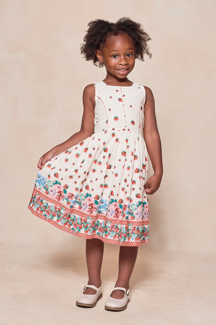 A young girl smiles in the Ivy City Co Mini Sadie Dress in Strawberry Vintage Tier—a sleeveless white cotton dress tiered with red strawberries and colorful florals at the hem. She stands on a neutral background, holding her dress and wearing white shoes.