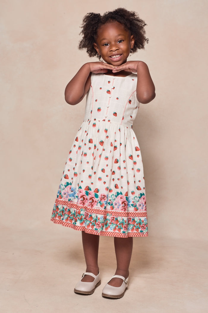 A young girl smiles with hands under her chin, wearing the Ivy City Co Mini Sadie Dress in Strawberry Vintage Tier—adorned with red dots and a colorful floral border—for a perfect mom and me matching look, paired with white shoes against a beige background.
