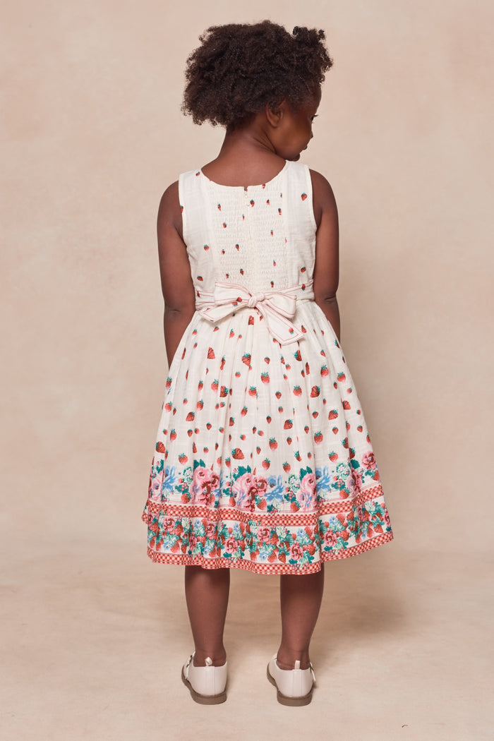 A young girl with curly hair faces away, wearing Ivy City Co's Mini Sadie Dress in Strawberry Vintage Tier, featuring red polka dots, waist bow, floral hem details, and white shoes on a beige background.