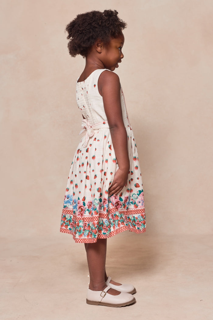 A young girl with curly hair stands sideways against a neutral background, wearing the Mini Sadie Dress in Strawberry Vintage Tier by Ivy City Co—a sleeveless, cream-colored cotton dress with tiers and colorful floral and polka dot patterns.