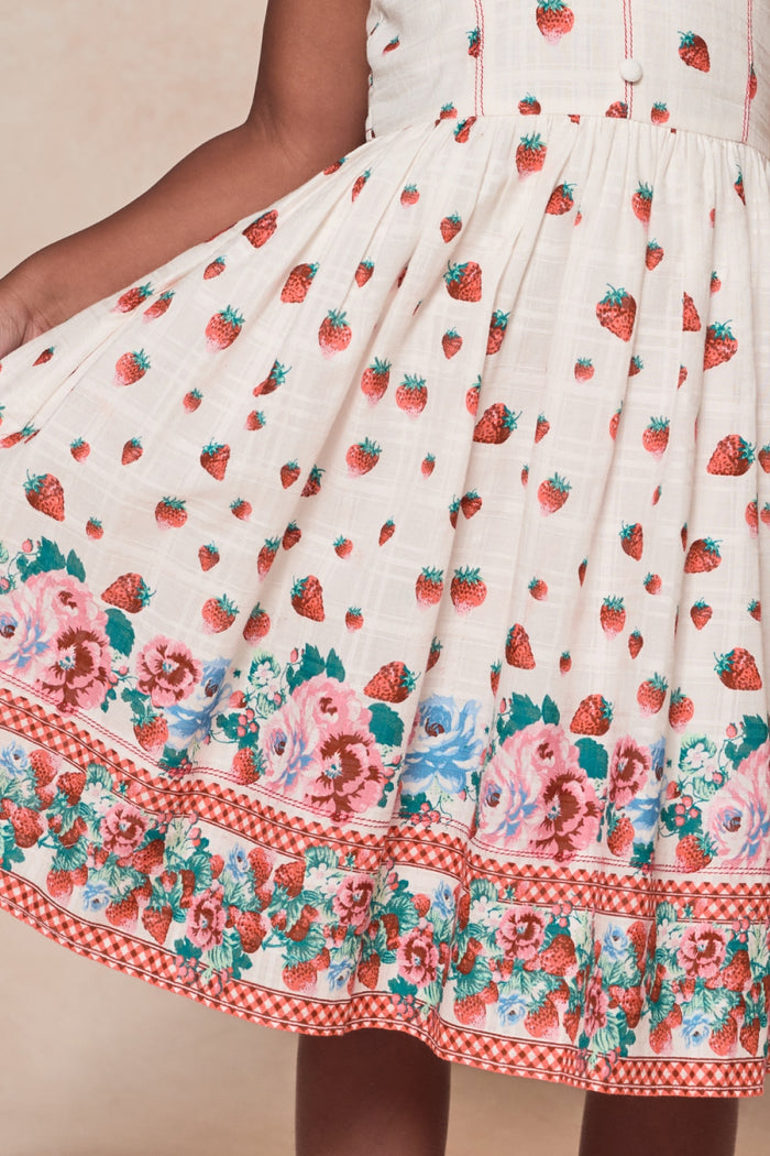 A person wearing the Ivy City Co Mini Sadie Dress in Strawberry Vintage Tier holds the skirt out, displaying its detailed print of red strawberries and pink and blue flowers along the hem.