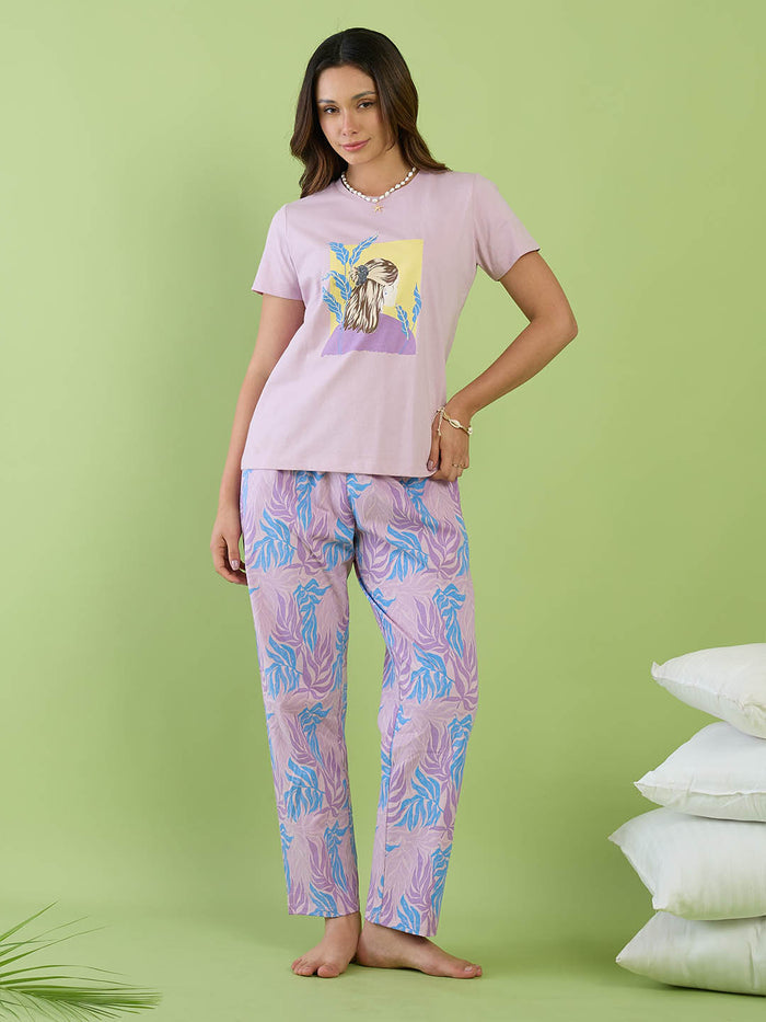 A woman modeling a two-piece pajama set consisting of a light pink t-shirt with a colorful graphic print and matching light pink pajama pants with a blue and purple palm leaf pattern, set against a solid light green background.
