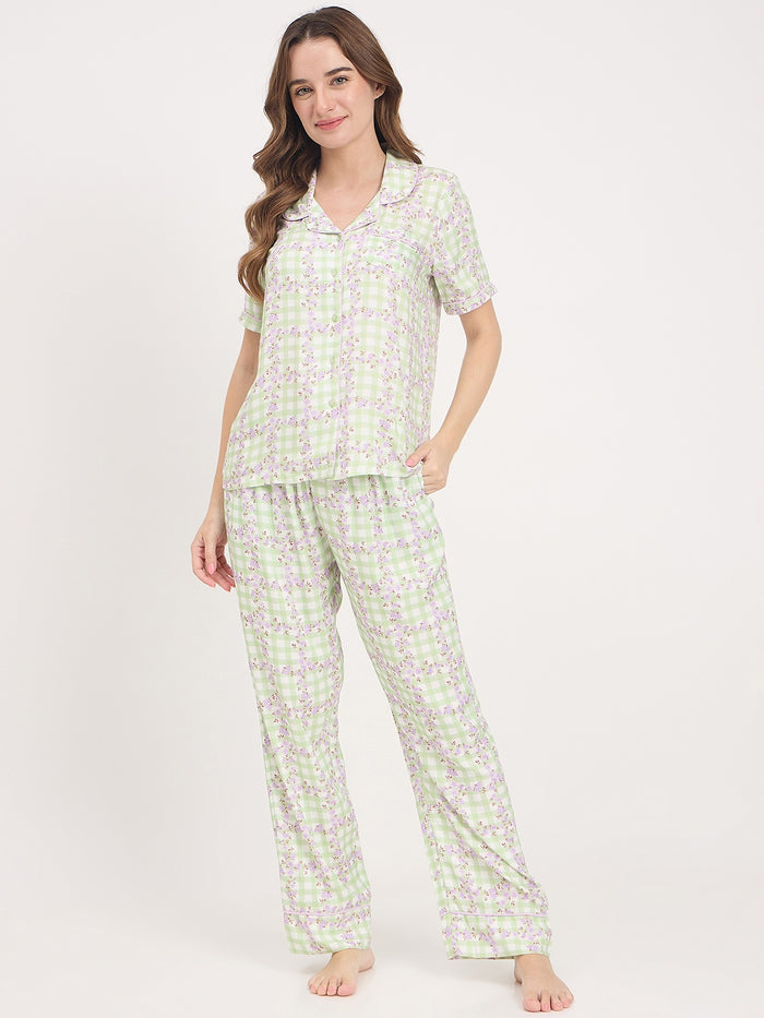 A woman modeling a two-piece pajama set with a short-sleeve collared top and matching wide-leg pants, featuring a light green and lavender floral pattern on a white background.