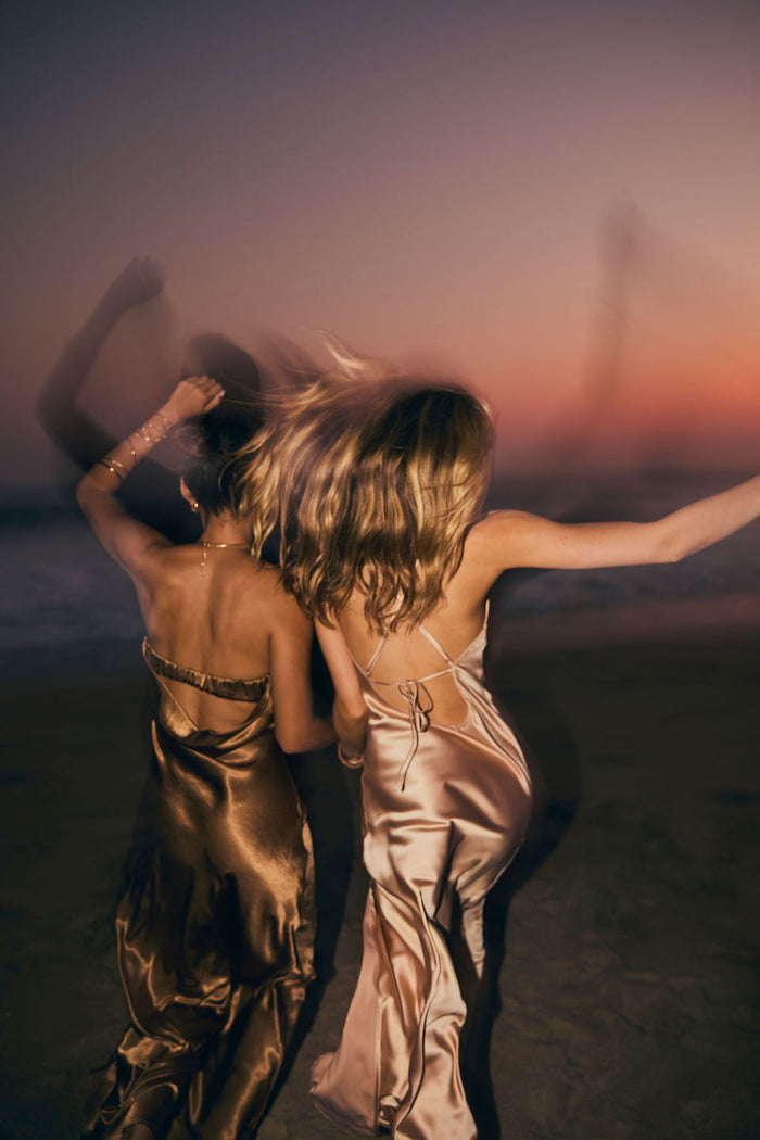 Back view of the bronze strapless satin maxi dress on the beach at sunset, beside a champagne slip dress.