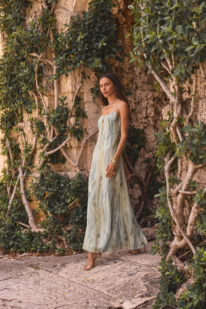 Sage watercolor chiffon maxi dress worn with gold sandals beside an ivy-covered stone wall.