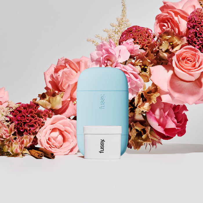 Fussy's refillable deodorant in Night Tales scent - a refillable deodorant case in Pale Blue and a refill pack - set against a backdrop of pink and red flowers.