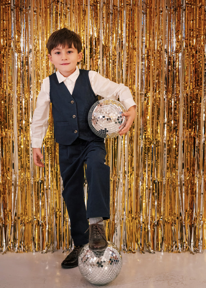 Boy wearing children's party clothes, dark blue pants and white shirt, standing in front of golden party curtain with disco ball