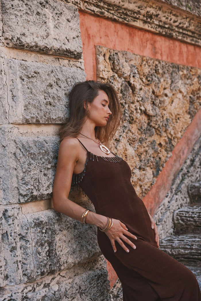 Espresso brown beaded maxi dress worn on-model, leaning against a coral stone wall.