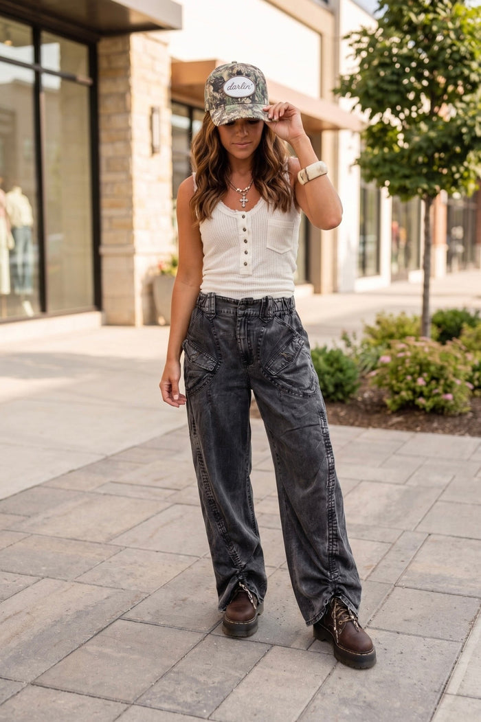 Boho western outfit with dark wash wide leg denim pants, white waffle knit tank top, camo trucker hat, and chunky lace-up boots styled for an edgy everyday boutique look.