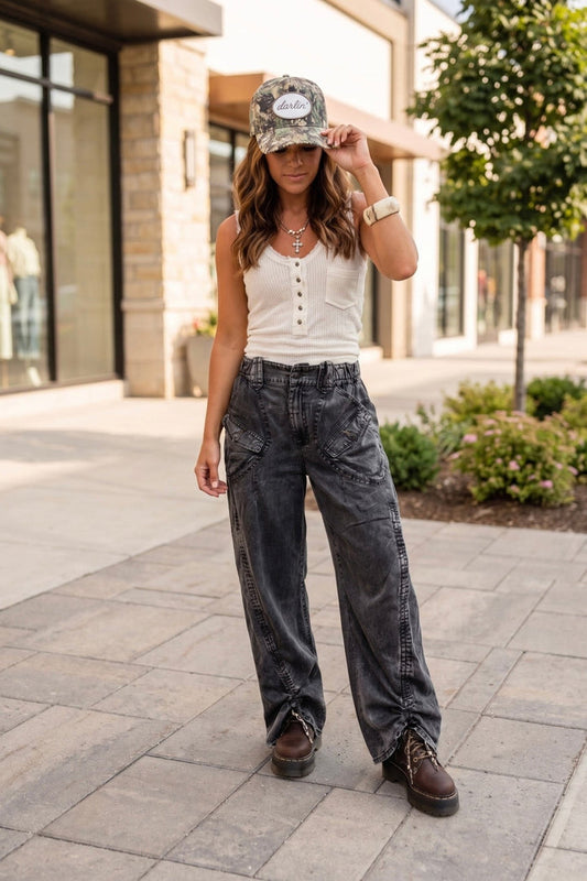 Boho western outfit with dark wash wide leg denim pants, white waffle knit tank top, camo trucker hat, and chunky lace-up boots styled for an edgy everyday boutique look.